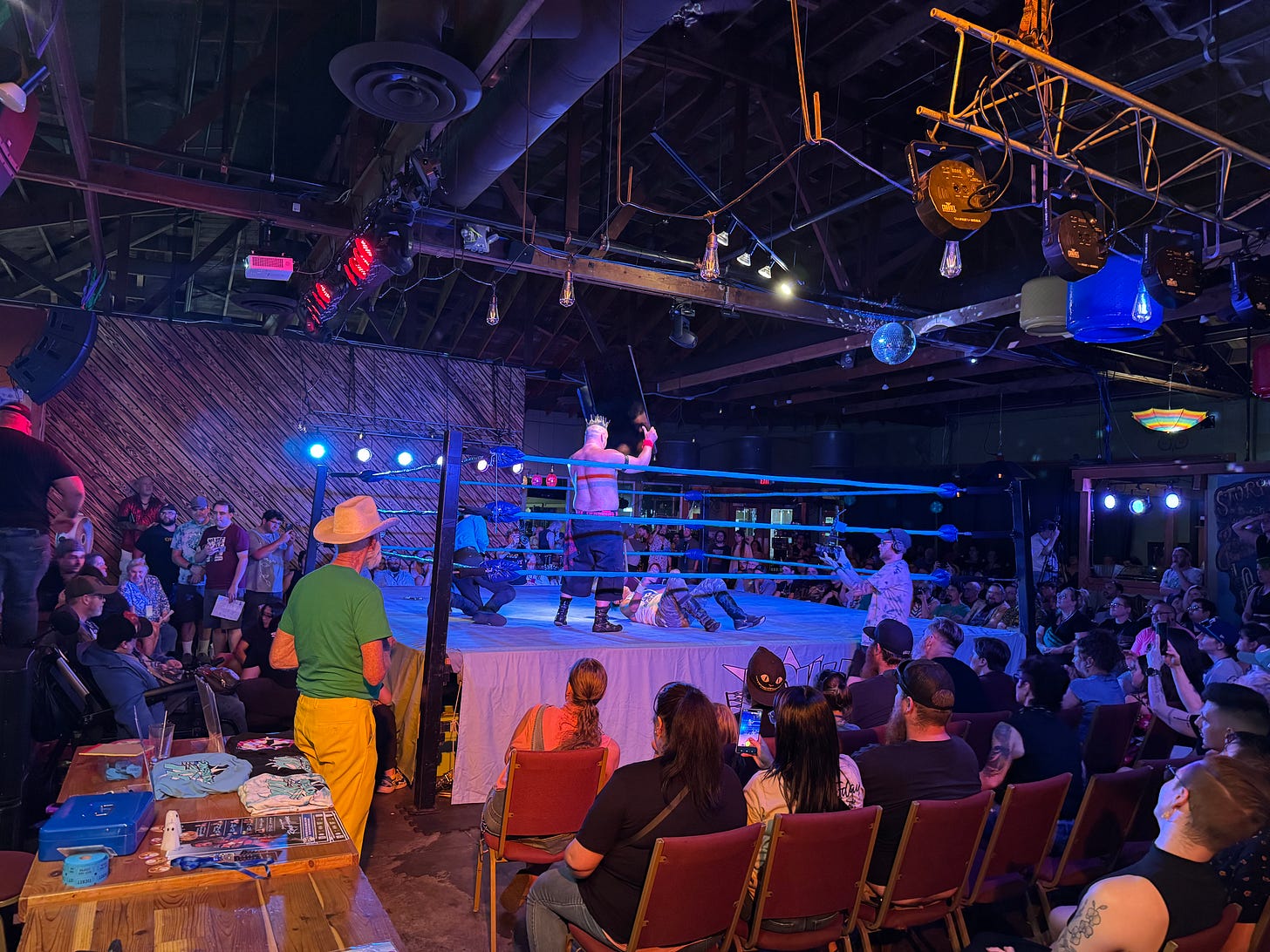 Indoor wrestling ring surrounded by audience in folding chairs, with exposed ceiling, neon lighting, and a man in a green shirt and cowboy hat watching from ringside