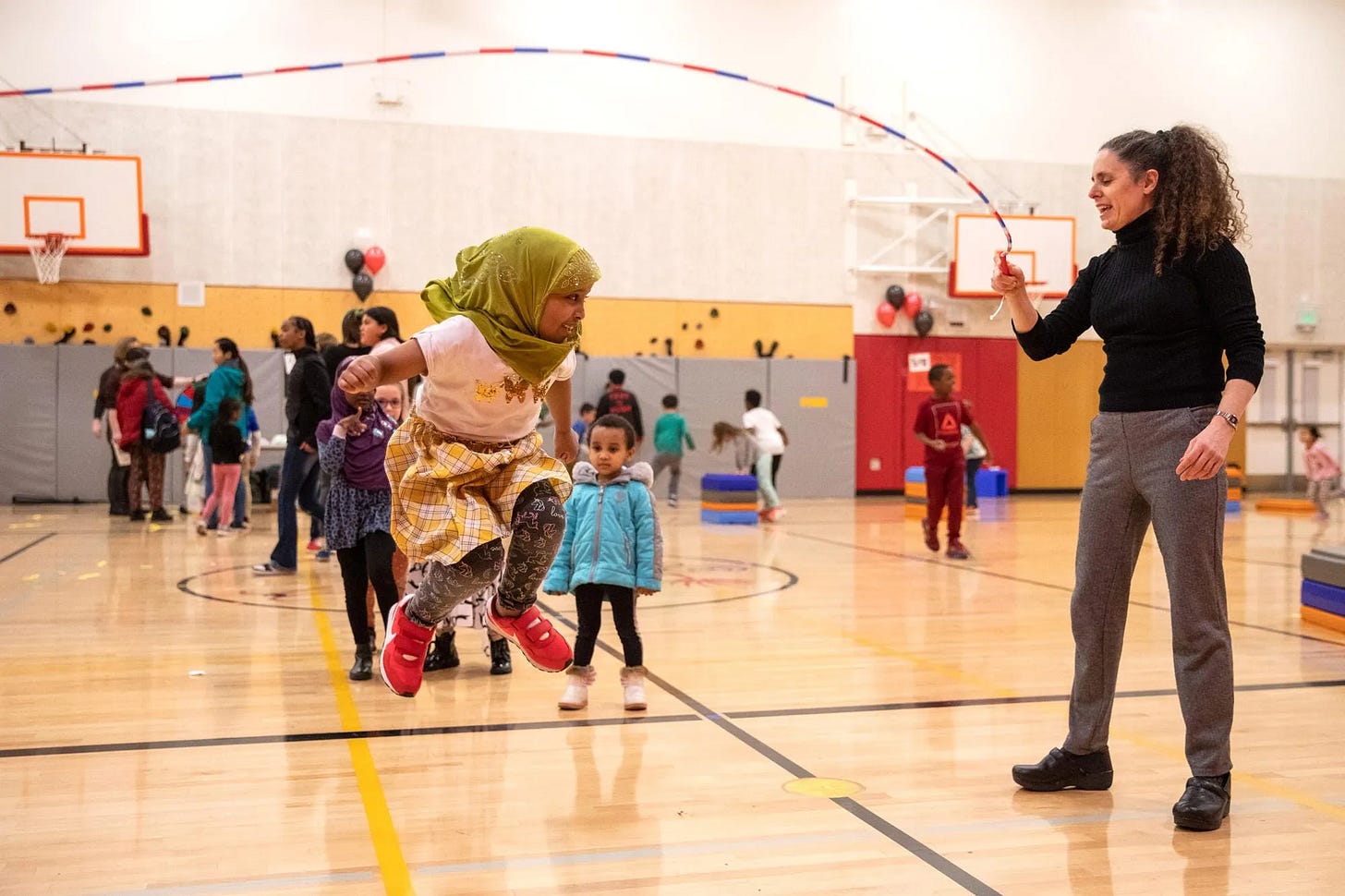 Photo of a school gym full of kids and adults. In the foreground, a white adult woman with curly hair swings one end of a jumprope, and in the middle of the frame, a smiling child wearing a green hijab and red sneakers is caught mid-jump.