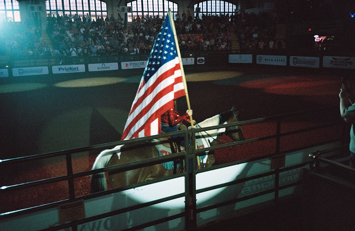 A woman in a sparkly red shirt is riding a brown horse in a rodeo arena, carrying a large American Flag with a single spotlight on her. Photograph taken by Connor Fowler on 35mm film in Fort Worth, Texas.