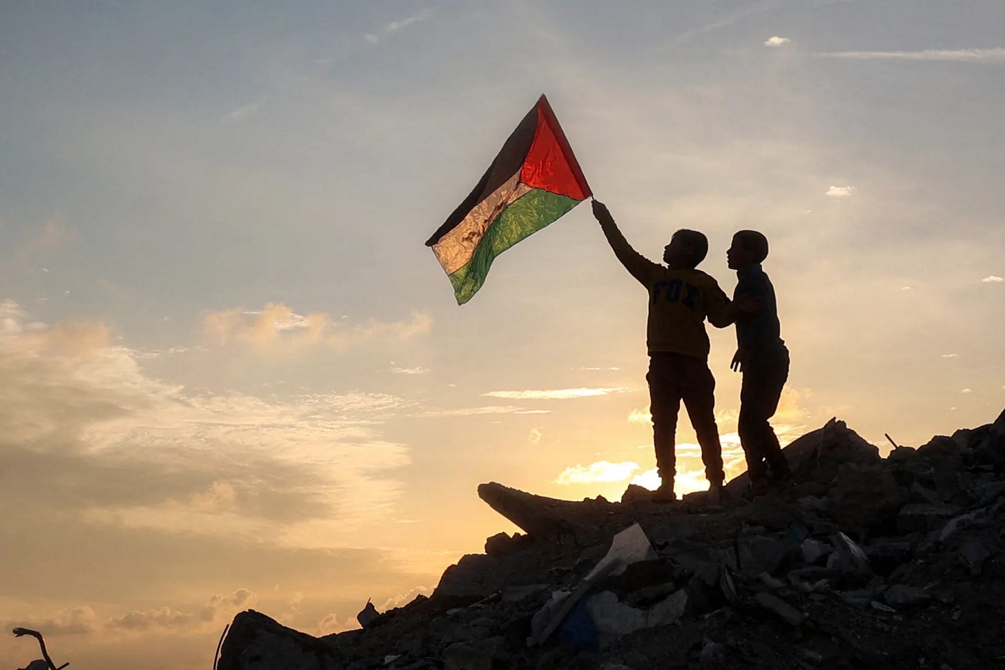 two little boys standing on a hill of rubble, holding up a palestinian flag against a sunset backdrop