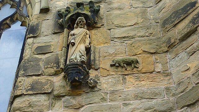 A carved statue of a man stands in a wall niche, looking down at a carved pig on the wall of a church tower.