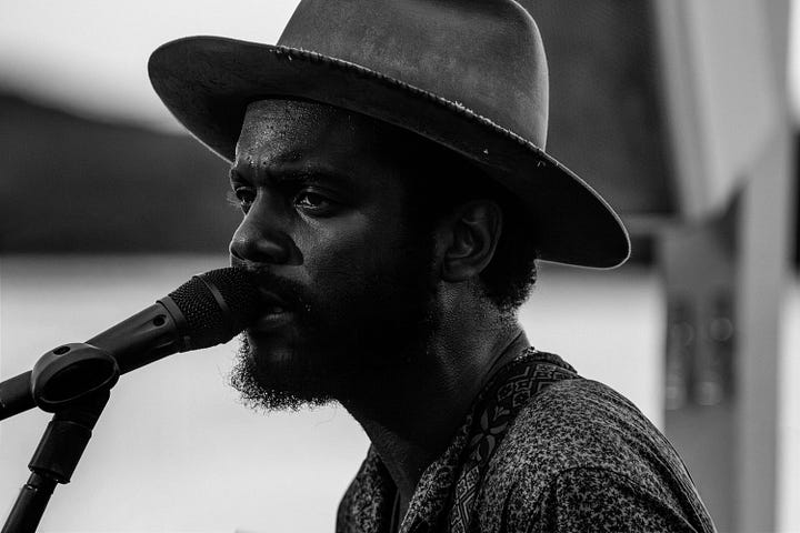 A collage of five photos of Gary Clark Jr. performing outdoors. The images are a mix of color and black and white, featuring close-ups of his face as he sings and wider shots of him playing the guitar, all while wearing his signature hat.