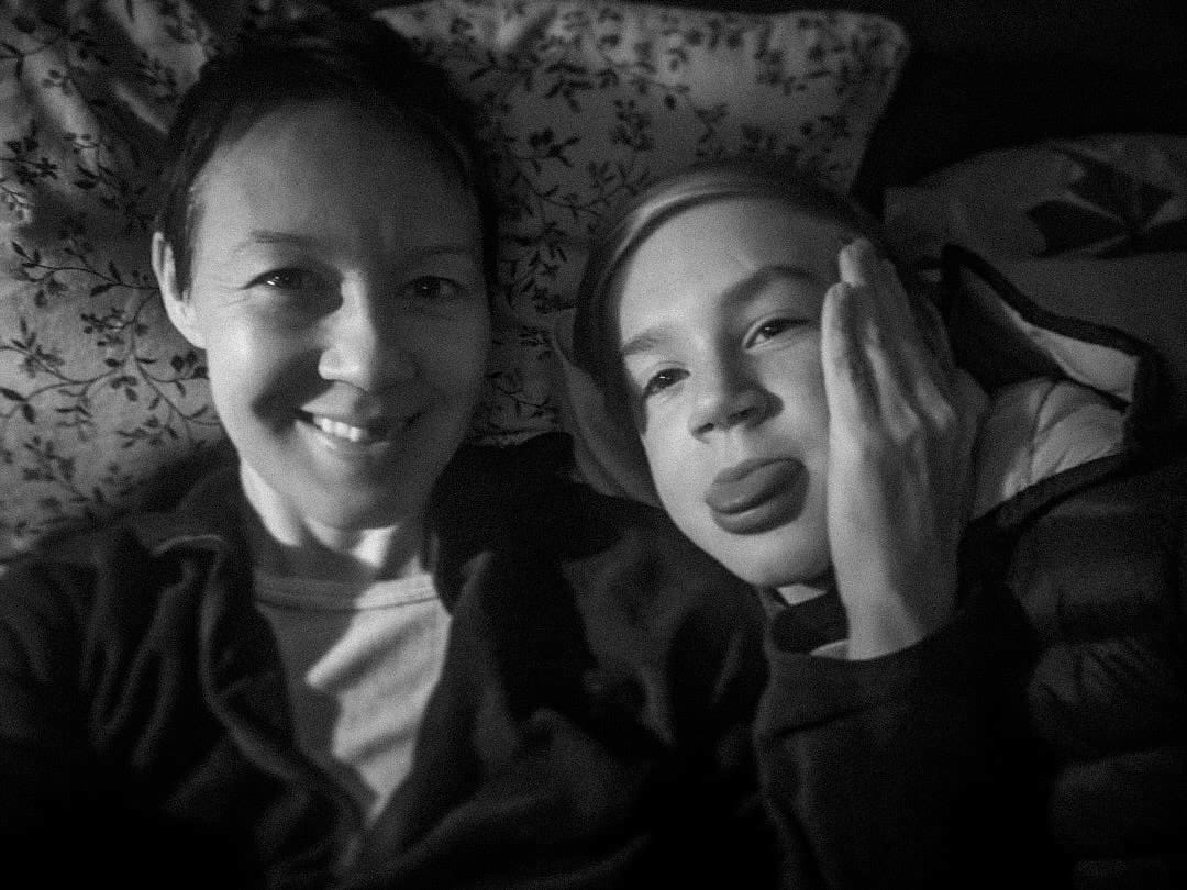4. Black and white photograph of Yvie a white woman with cropped brown hair, smiling at the camera, laying in bed against a pillow. Her son is on her left also looking at the camera, pulling his tongue. Her left hand rests against his face. 4. Black and white photograph of Yvie a white woman with cropped brown hair, smiling at the camera, laying in bed against a pillow. Her son is on her left also looking at the camera, pulling his tongue. Her left hand rests against his face.