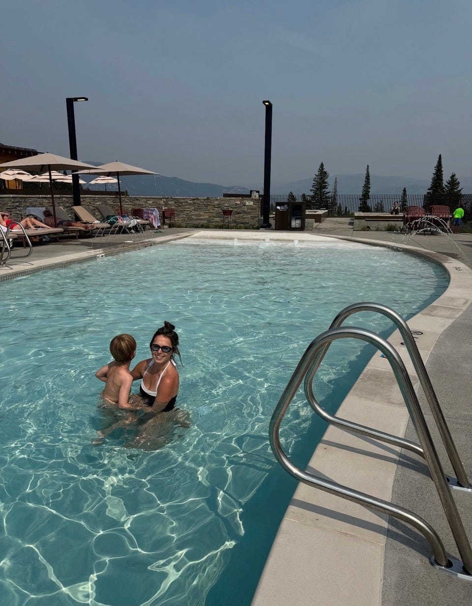 A woman holds her young child in a public swimming pool. In the distance are trees and mountains.