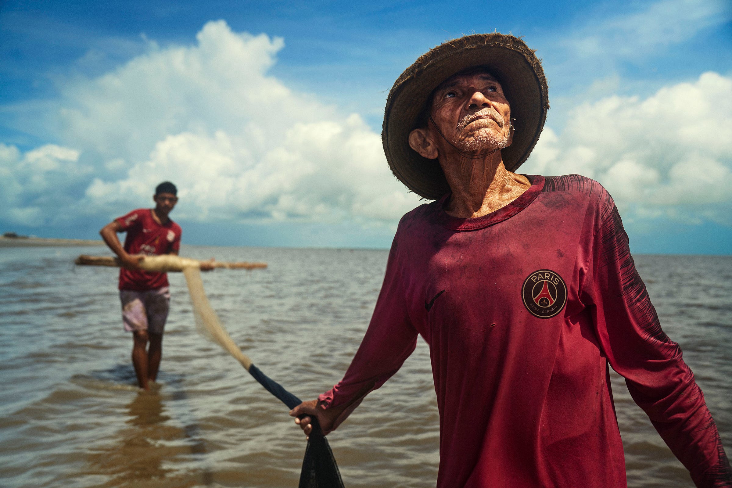 Two men stand in shallow water under a blue sky with clouds. The older man in front wears a wide-brimmed hat and a maroon Paris Saint-Germain shirt, while the man behind him pulls a fishing net.