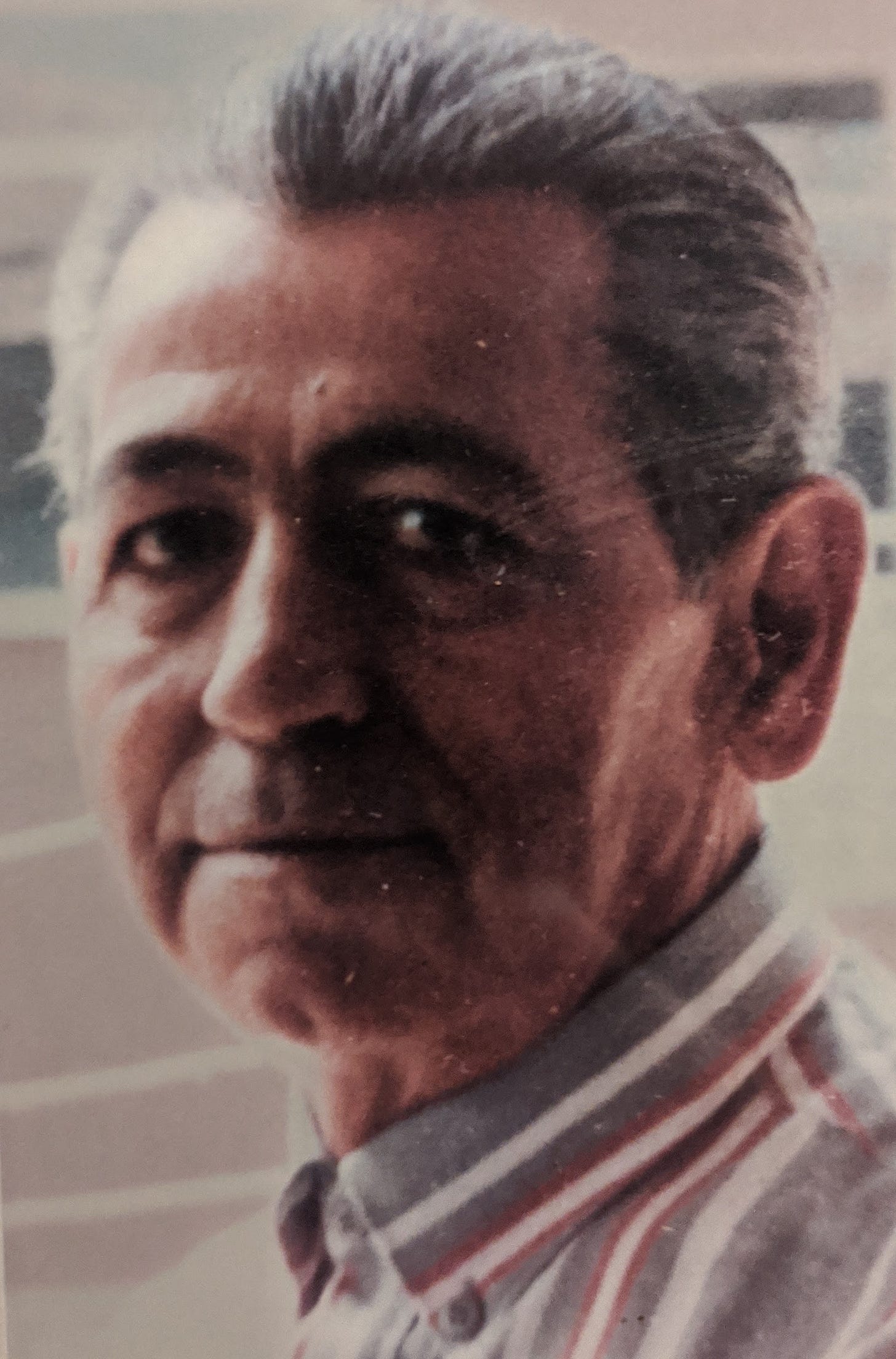 60 yaşlarında bir erkek. Yüzünde değişik, özgüvenli, hafif hüzünlü bir bakış. Kır saçları arkaya taranmış. Bir gömlek giymiş.