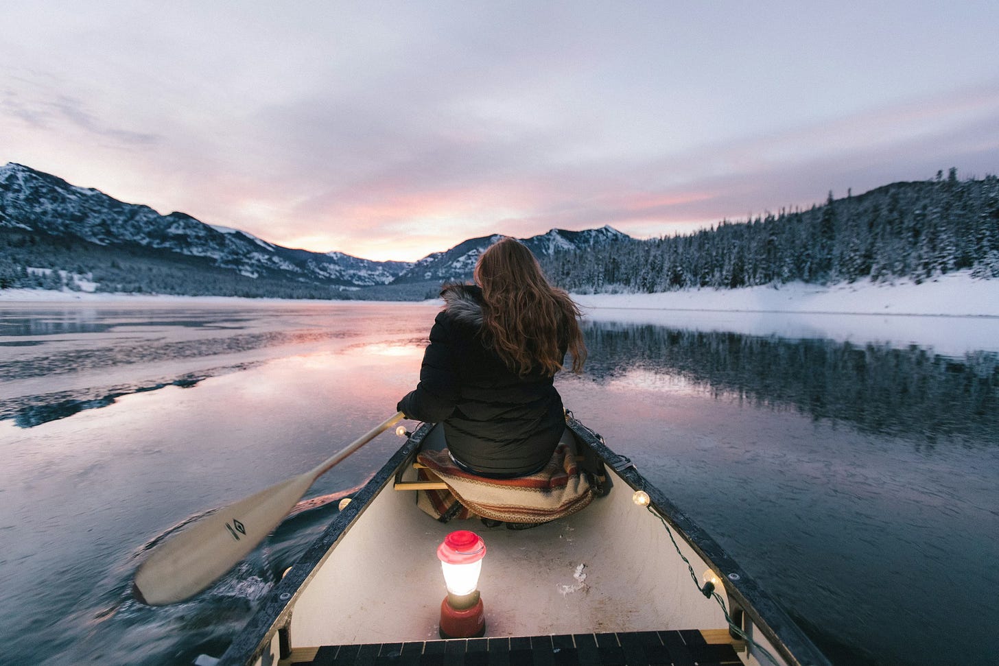 A woman in the front of a canoe paddles in a lake surrounded my snow and hills at sunrise.