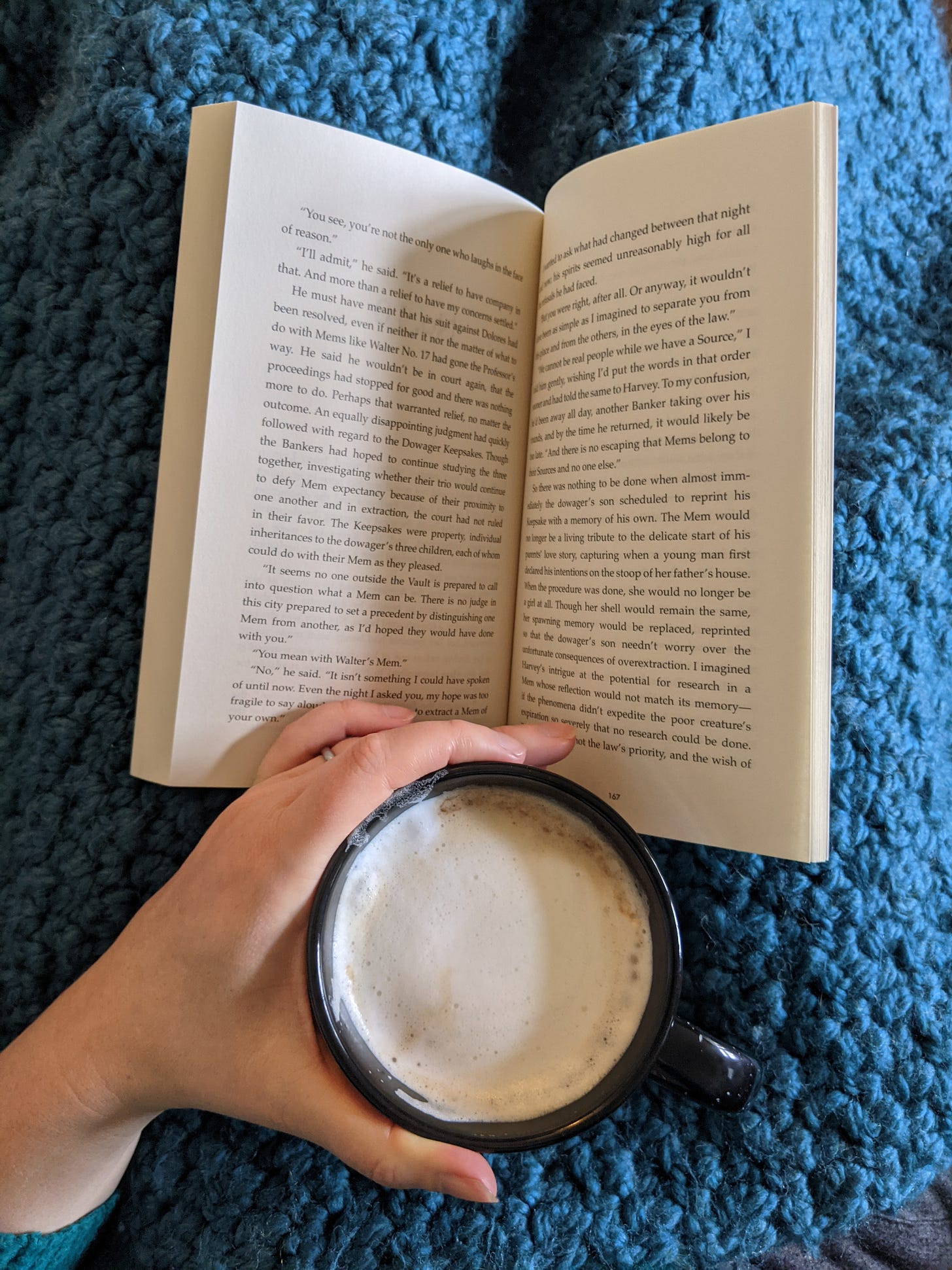 an open book on a blue blanket with a hand holding a cup of coffee