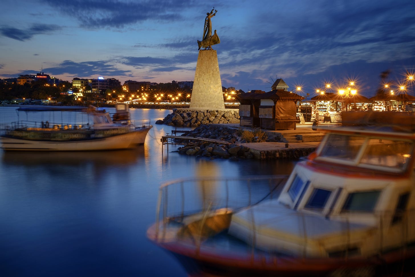 Boats bobbing around in the harbour at dusk. A statue of a person stands on a stone monument.