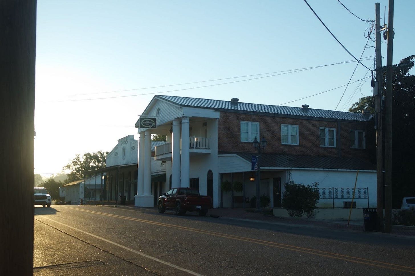 A brick building with white columns and a metal roof sits along a street with a parked red vehicle in front, utility poles visible overhead.