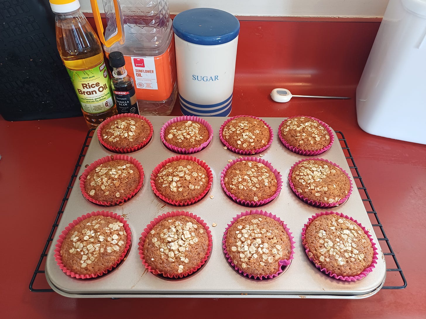 A tray of delightfully brown muffins in a tray, sitting on a rack set on a red bench. In the background are some of the ingredients, looking suspiciously neat, and also a blue and white cannister labelled "SUGAR". This is a lie. That's where I keep my salt. A tray of delightfully brown muffins in a tray, sitting on a rack set on a red bench. In the background are some of the ingredients, looking suspiciously neat, and also a blue and white cannister labelled "SUGAR". This is a lie. That's where I keep my salt.