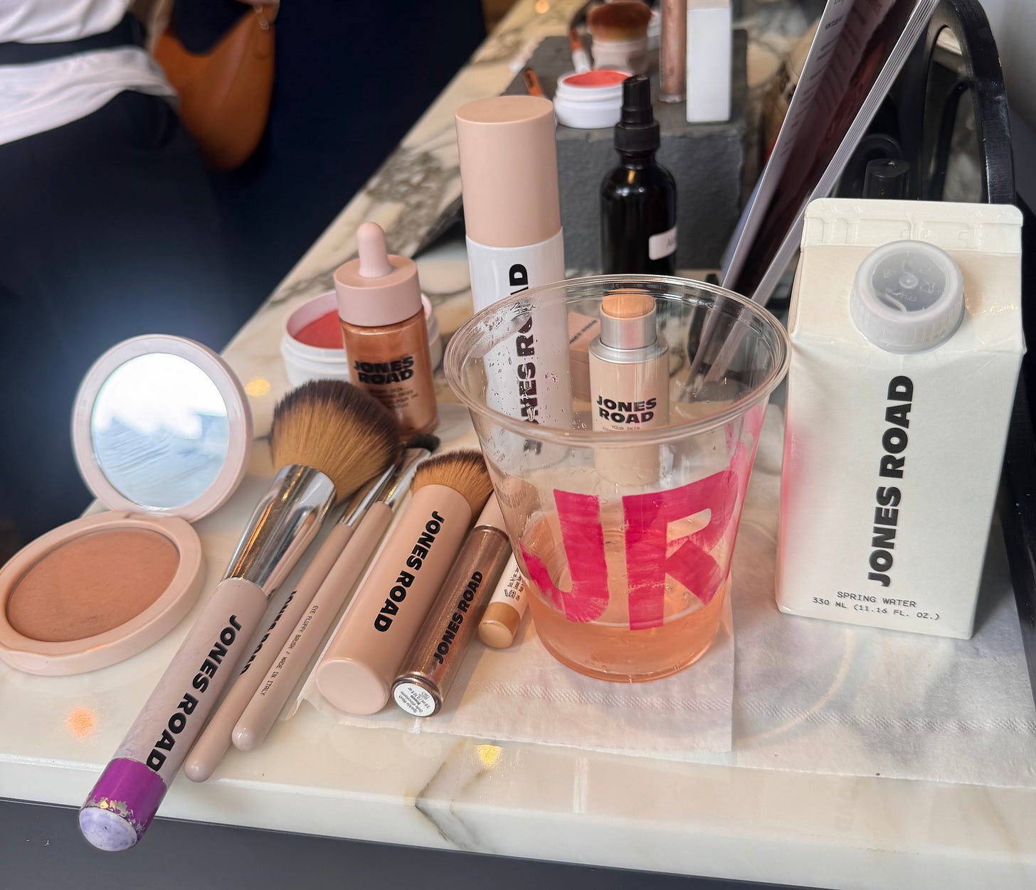 A flat lay photograph taken from above on a marble surface, showcasing a collection of Jones Road beauty products and makeup brushes. On the left, an open compact reveals a bronzer next to a sleek pink-cased mirror and two professional makeup brushes. In the center, a clear plastic cup branded with pink "JR" lettering holds a pink beverage with a straw, surrounded by a pump bottle of Bright Skin Illuminating Drops, a foundation stick, and a slender concealer tube. To the right, a white Jones Road-branded paper carton of spring water sits next to a purple-handled eye blending brush. A flat lay photograph taken from above on a marble surface, showcasing a collection of Jones Road beauty products and makeup brushes. On the left, an open compact reveals a bronzer next to a sleek pink-cased mirror and two professional makeup brushes. In the center, a clear plastic cup branded with pink "JR" lettering holds a pink beverage with a straw, surrounded by a pump bottle of Bright Skin Illuminating Drops, a foundation stick, and a slender concealer tube. To the right, a white Jones Road-branded paper carton of spring water sits next to a purple-handled eye blending brush.