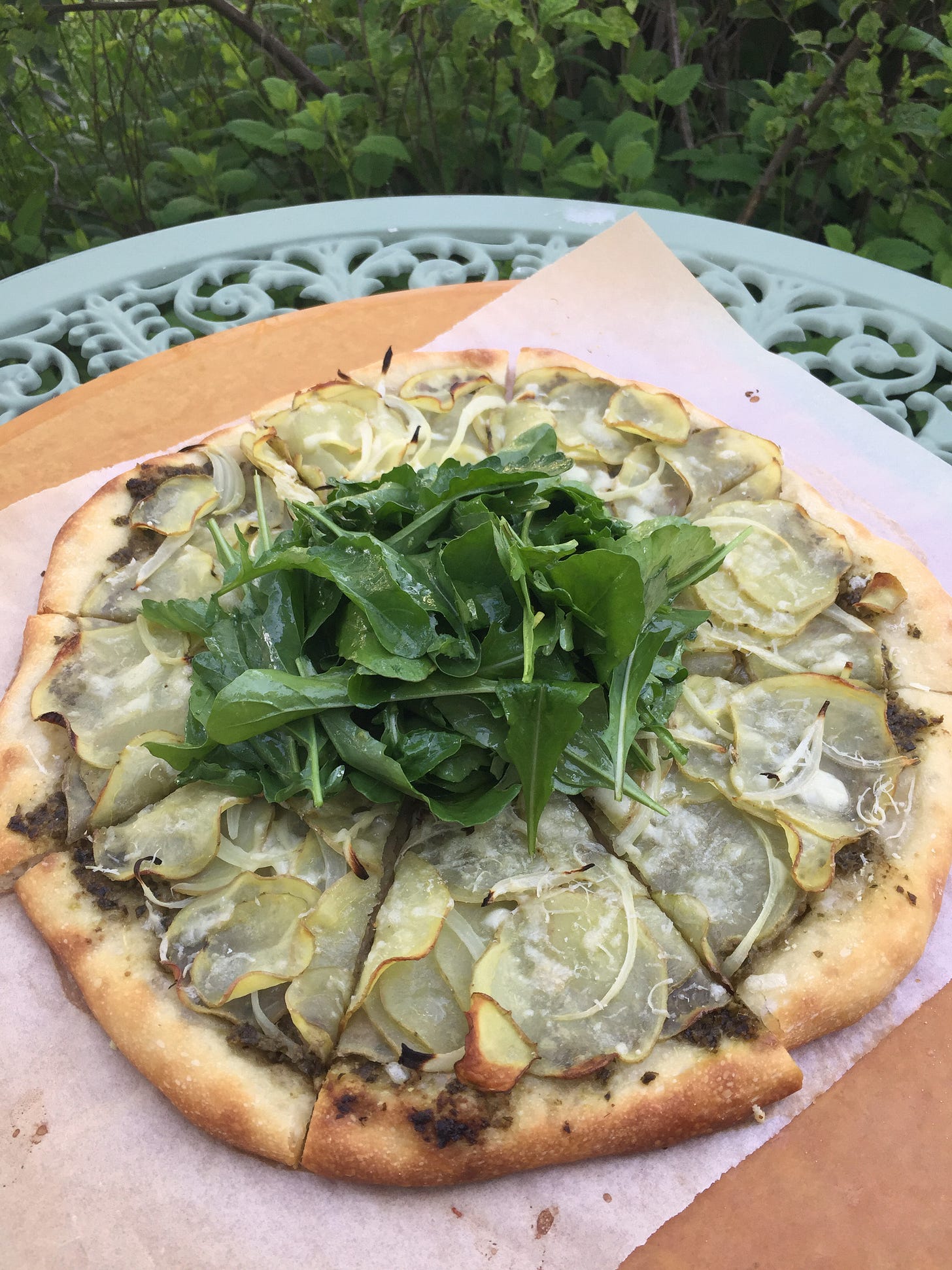 On a mint green wrought-iron table is a wooden pizza peel with a potato pizza on a piece of parchment. In the centre of the pizza is a large pile of arugula salad. On a mint green wrought-iron table is a wooden pizza peel with a potato pizza on a piece of parchment. In the centre of the pizza is a large pile of arugula salad.