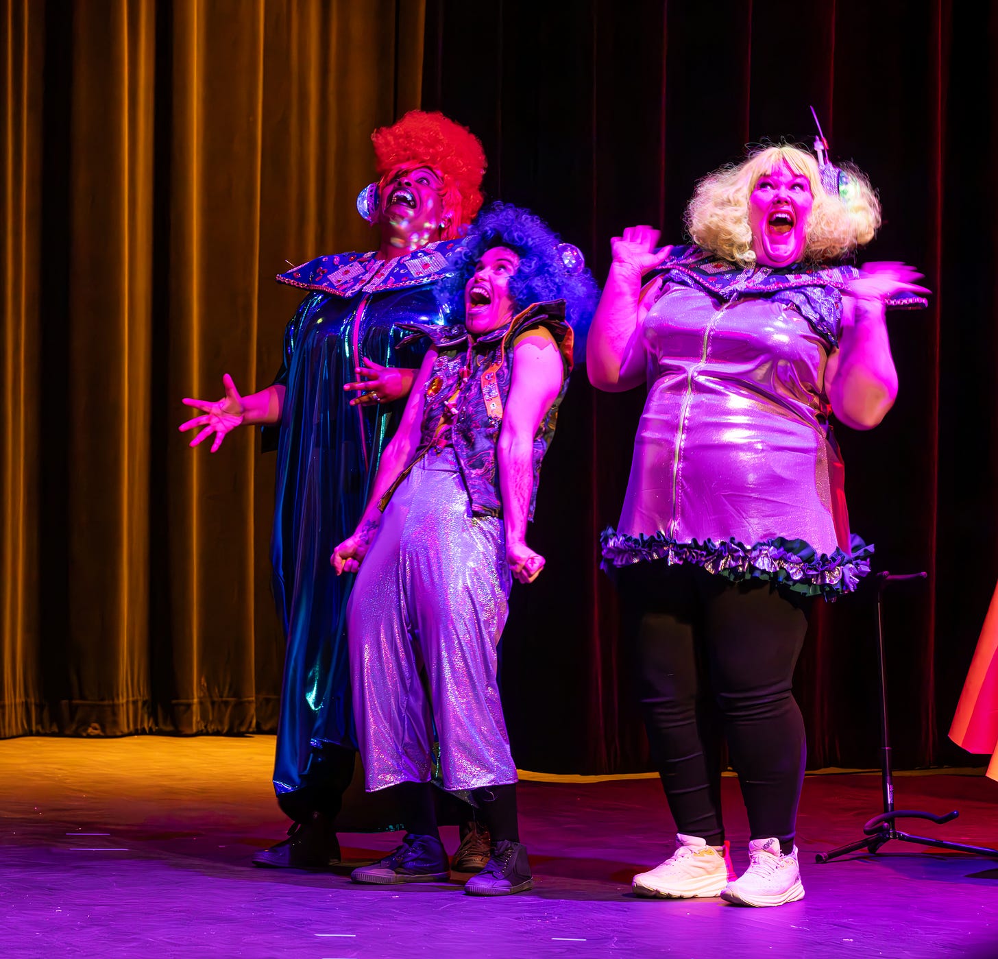 3 actors on stage dressed in brightly colored alien costumes laugh loudly, with their whole bodies