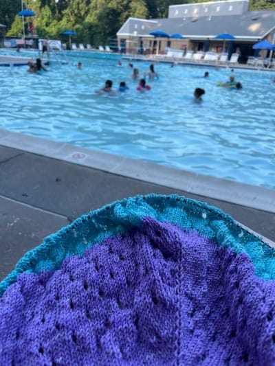 A swimming pool with children in the background. A partially-knit project in blue and purple is in the foreground