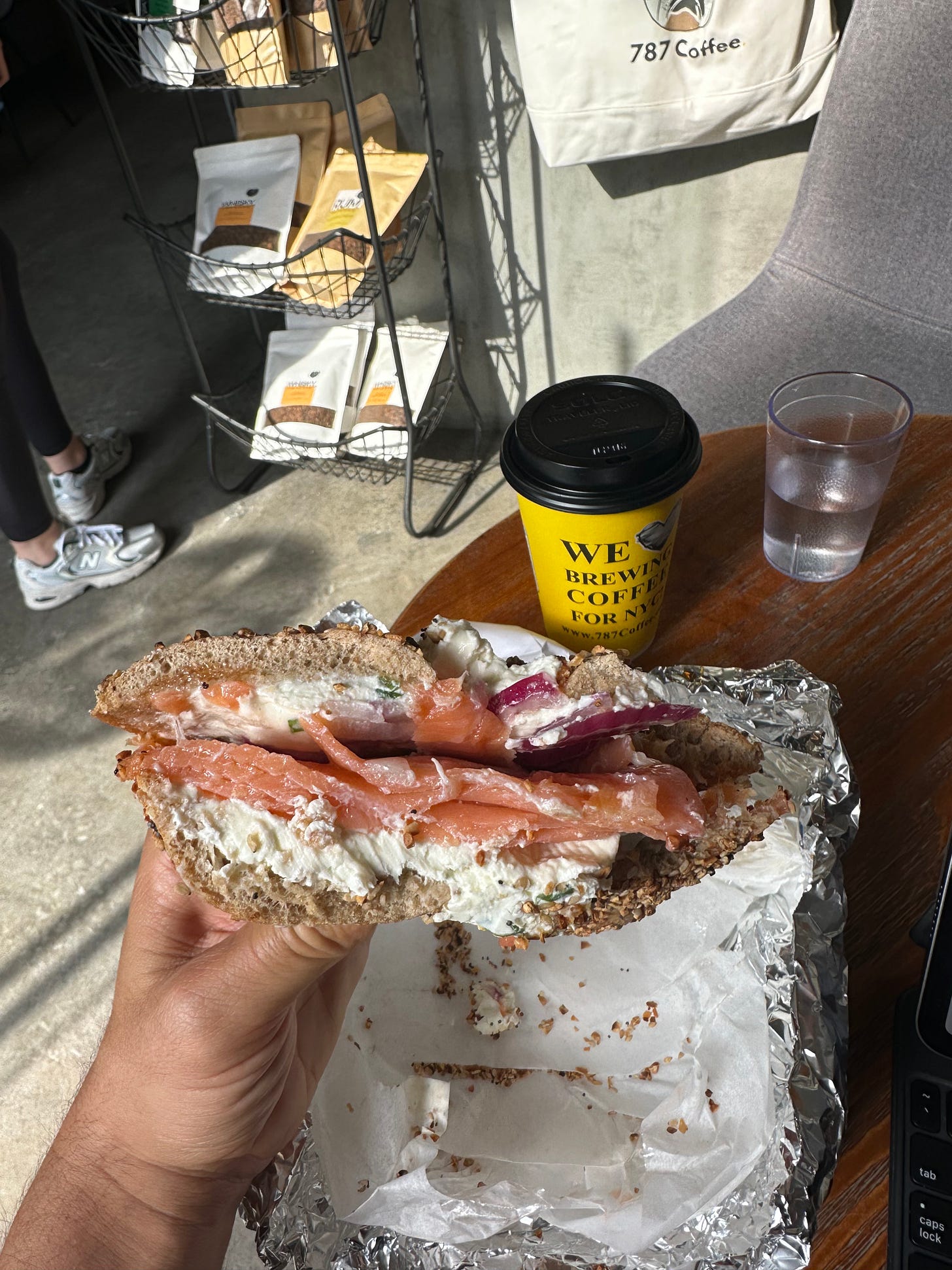 Jonathan is holding an everything flagel with cream cheese and lox. Behind the bagel is a cup of coffee in a yellow cup with black lids, and next to that a plastic cup of water.