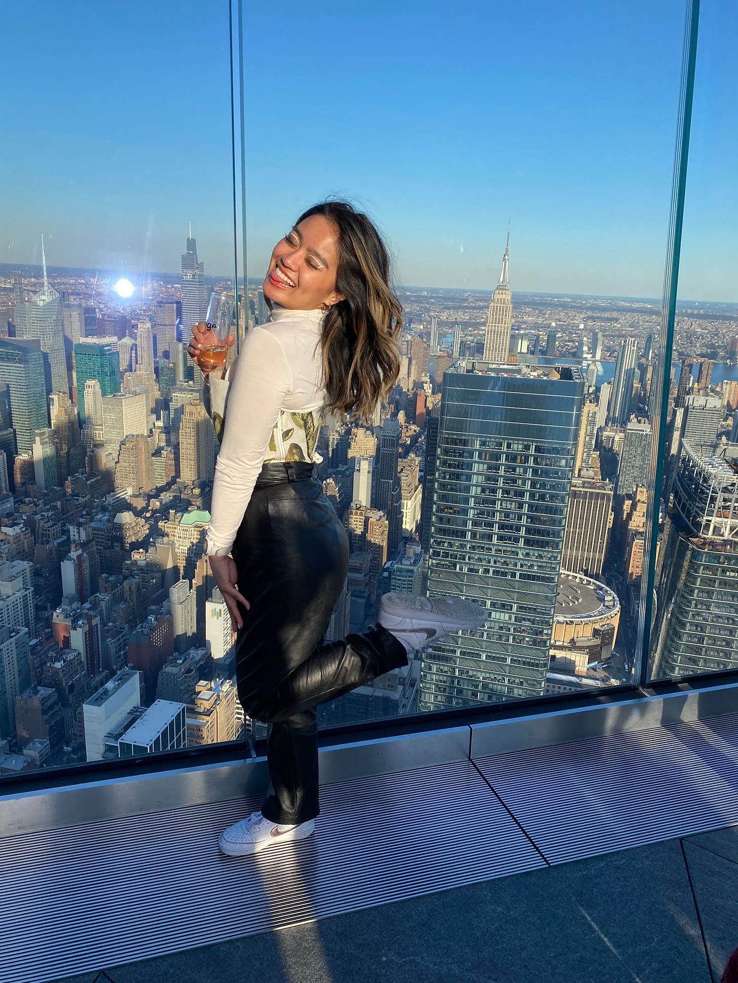 Joelle posing at the observation deck at The Edge in New York City with the Empire State Building in the background. She wears a white turtleneck top underneath a white corset with a green leaf pattern and black pleather pants. She's holding a champagne flute and is standing to the side with her leg popped back, eyes closed and smiling