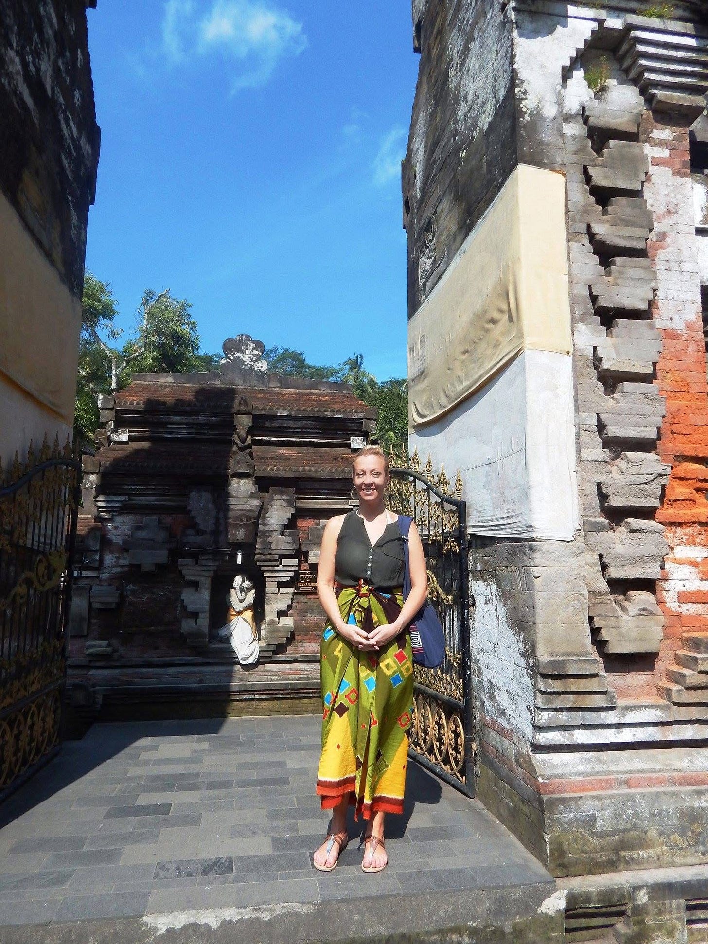 A woman standing at a temple entrance in Bali wearing a sarong, smiling in bright sunlight, surrounded by stone architecture.