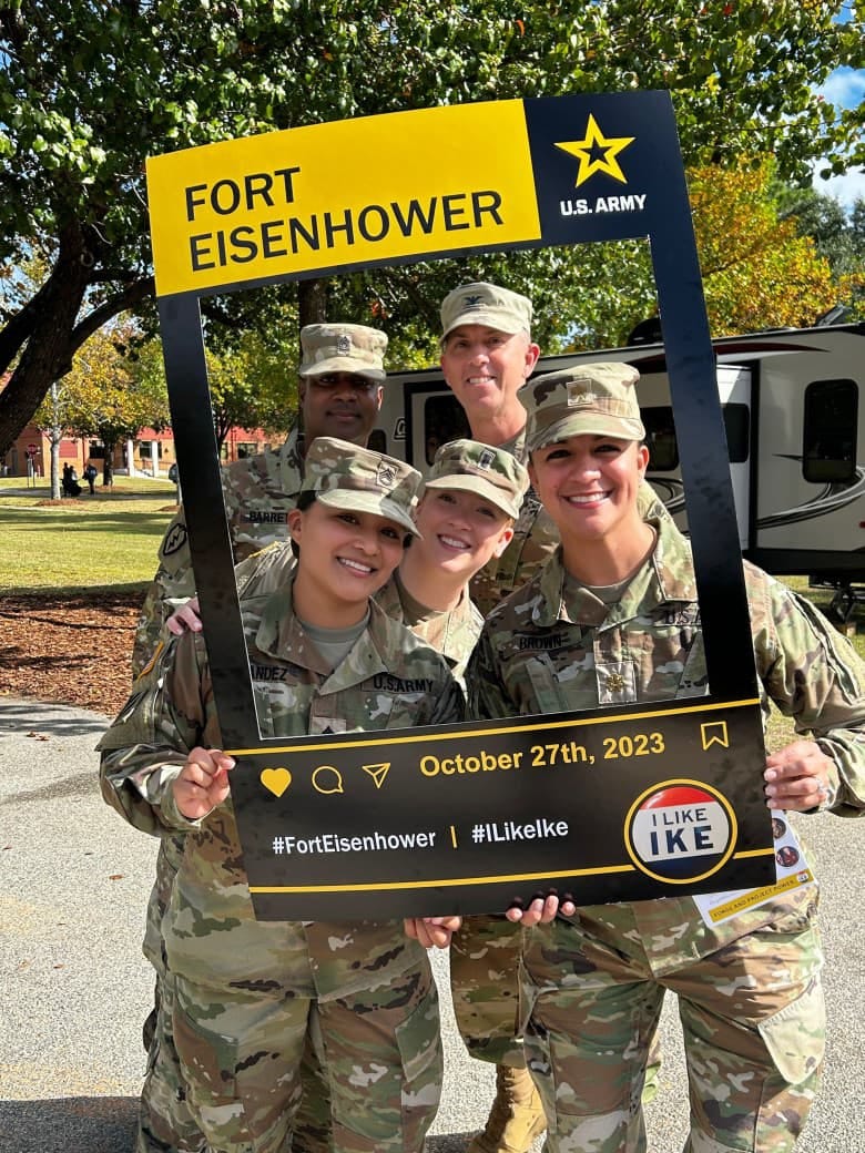 Soldiers on a sunny day hold up a yellow and black cardboard frame through which you can see their faces that shows support for Fort Eisenhower.
