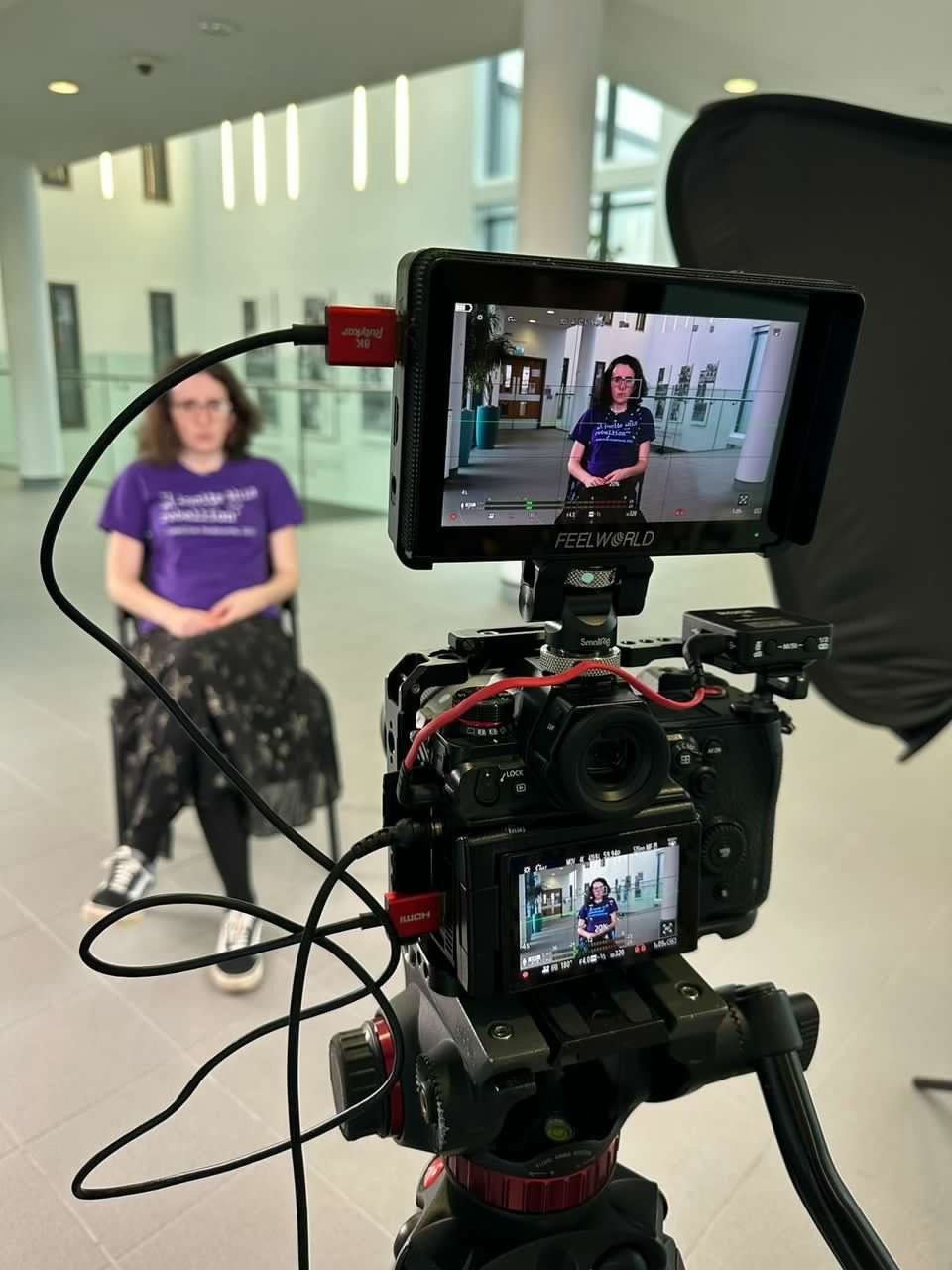 A white woman in a purple shirt and black skirt speaks to a camera while filming on location 