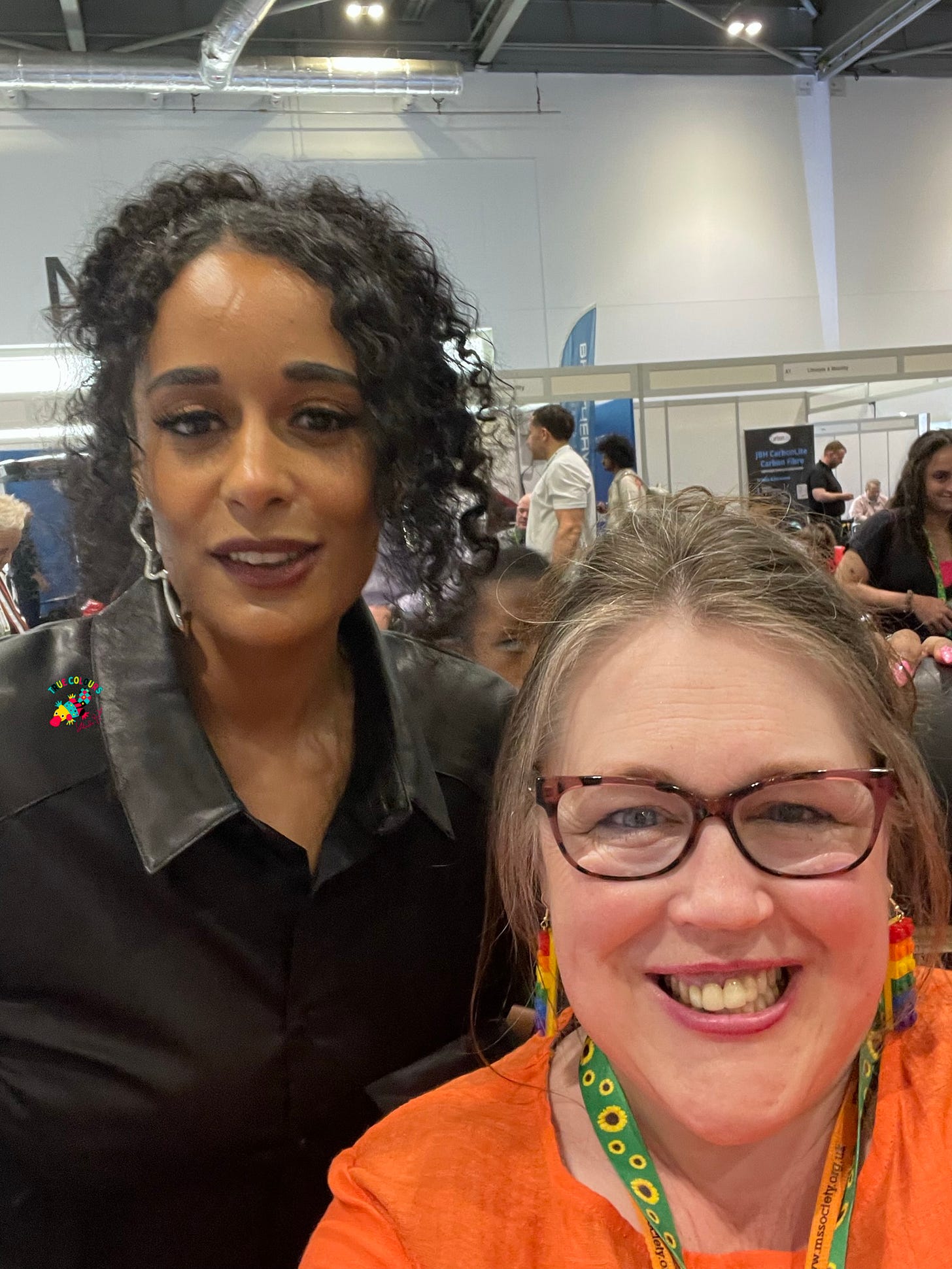 Roxy Murray is a lady who is standing.  She has brown skin and curly dark hair, Yolanda is in a wheelchair and is wearing an orange top, glasses and her brown hair is tied up.