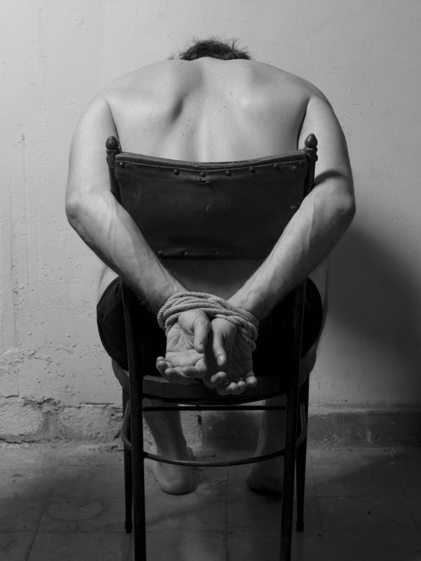 A black and white photo of a man tied to a chair. He's looking down and facing a concrete wall instead of facing us.