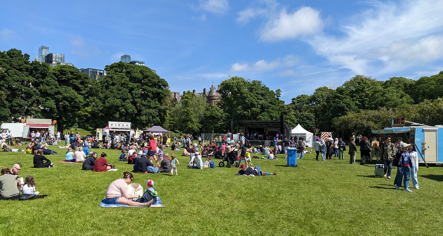 people sitting on the grass near a stage with food stalls in the background on a sunny day
