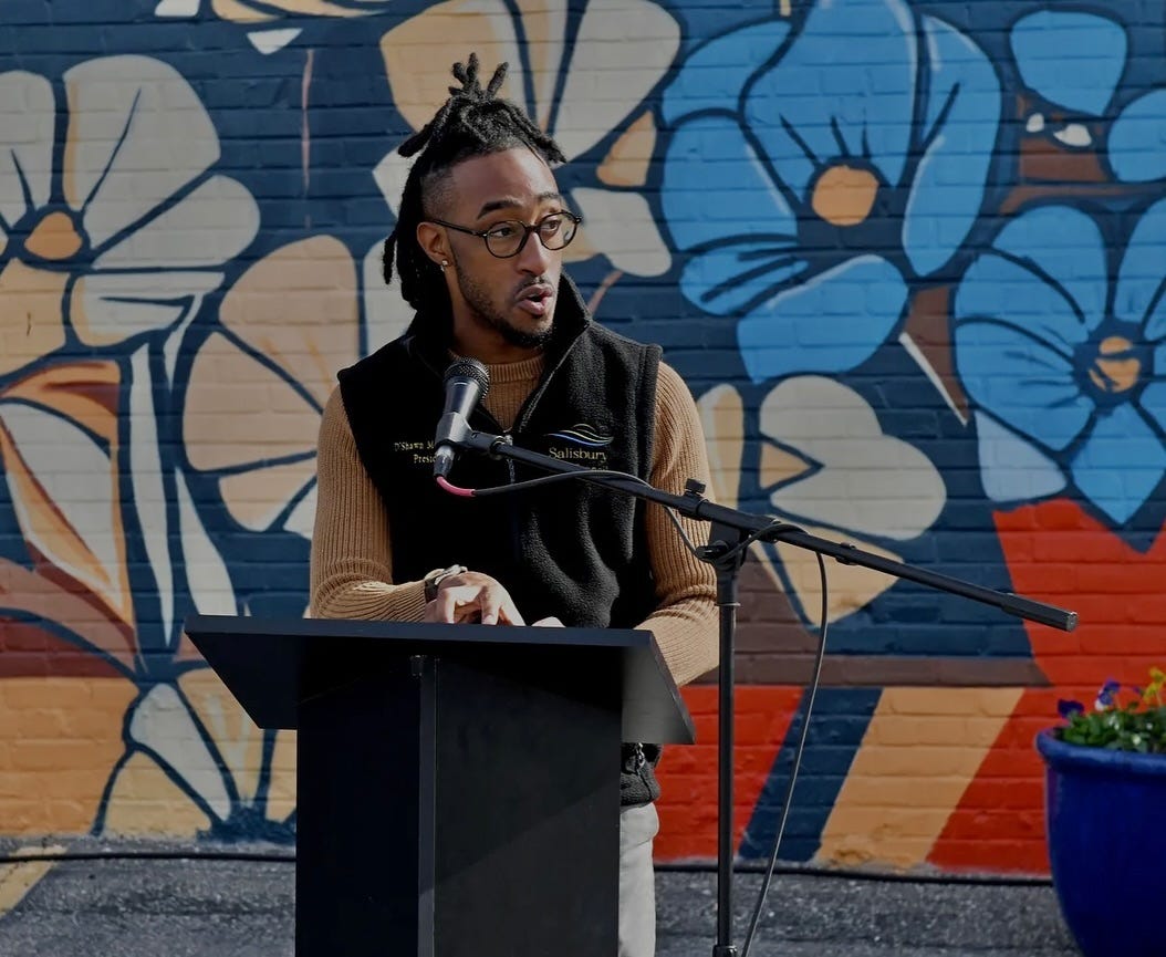 Doughty speaks at a podium, with a colorful mural in the background.