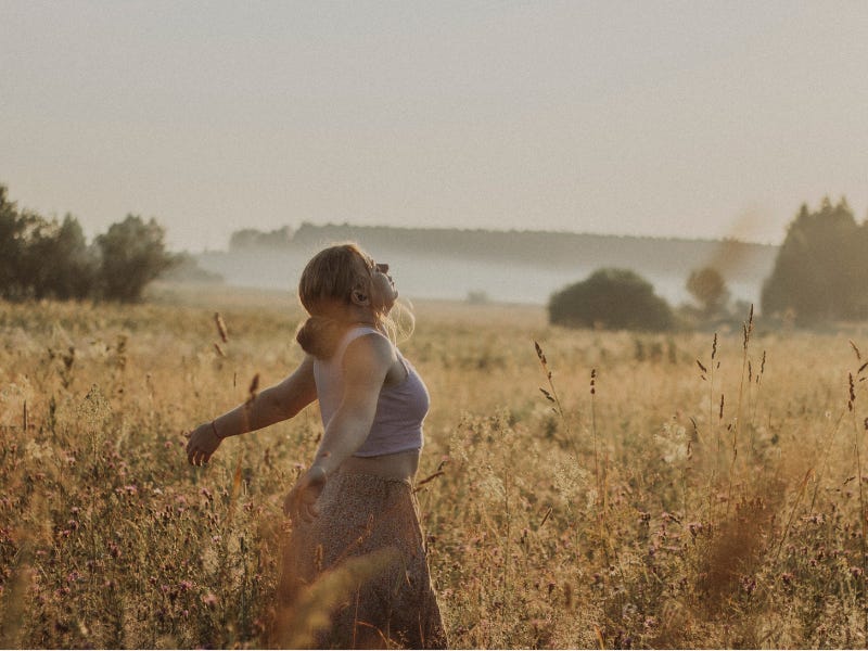 A woman with arms open standing in a sunny field with her eyes closed