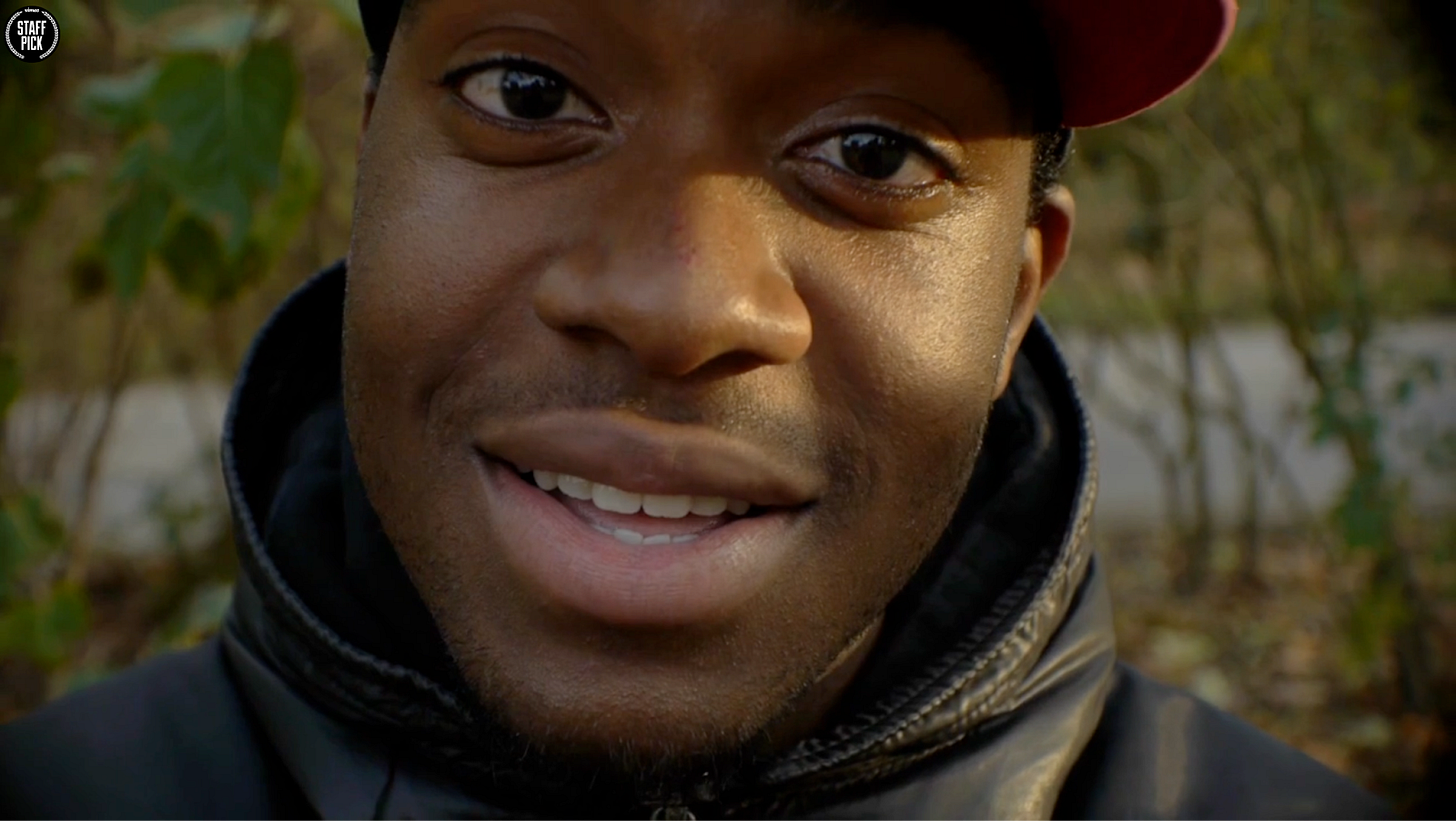 Close-up portrait of a smiling Black teenage boy wearing a red baseball cap and dark jacket, photographed outdoors with greenery in the background.
