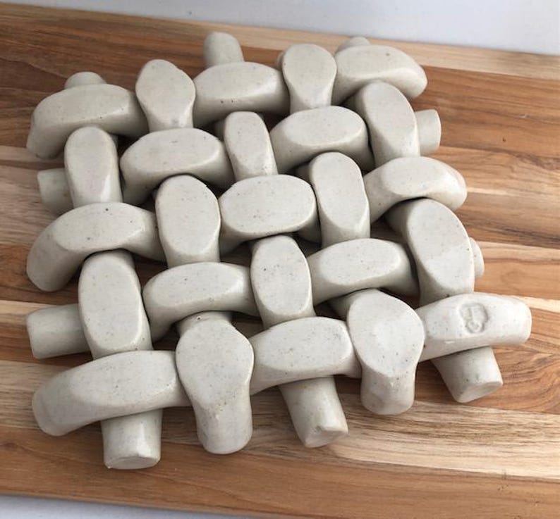 May include: A woven ceramic trivet with a light gray color and a speckled texture. The trivet is made of individual pieces that are woven together to form a square shape. The trivet has a small, circular mark on one of the pieces that reads '8'.