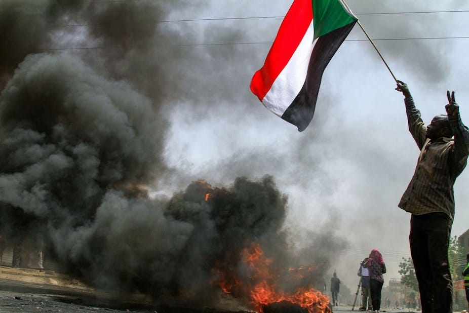 A man holding up a flag in one hand while the other hand shows the two-finger peace sign stands near heavy smoke created by a burning tyre. A man holding up a flag in one hand while the other hand shows the two-finger peace sign stands near heavy smoke created by a burning tyre.