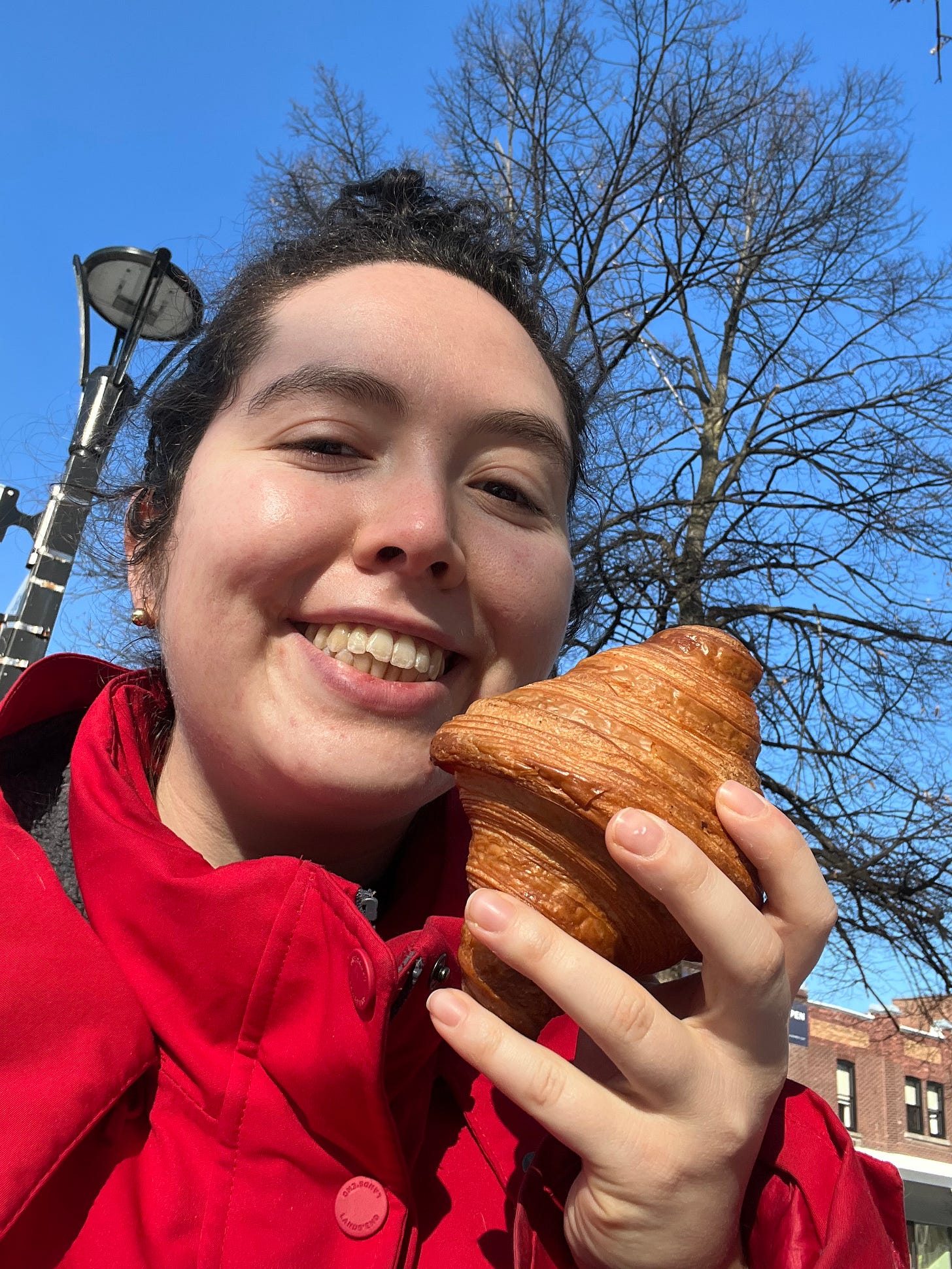 Shira, grinning, in a red coat, against a blue sky, holding a very golden blue croissant.