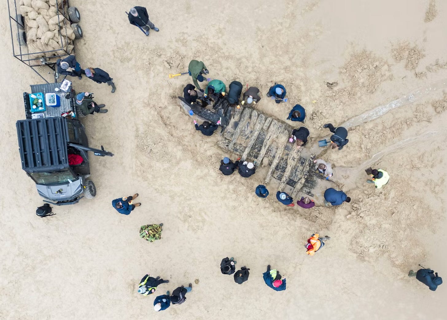 Aerial view of archaeologists gathered around the exposed wooden ribs of a 17th-century shipwreck on a sandy beach, with excavation tools and a utility vehicle nearby.