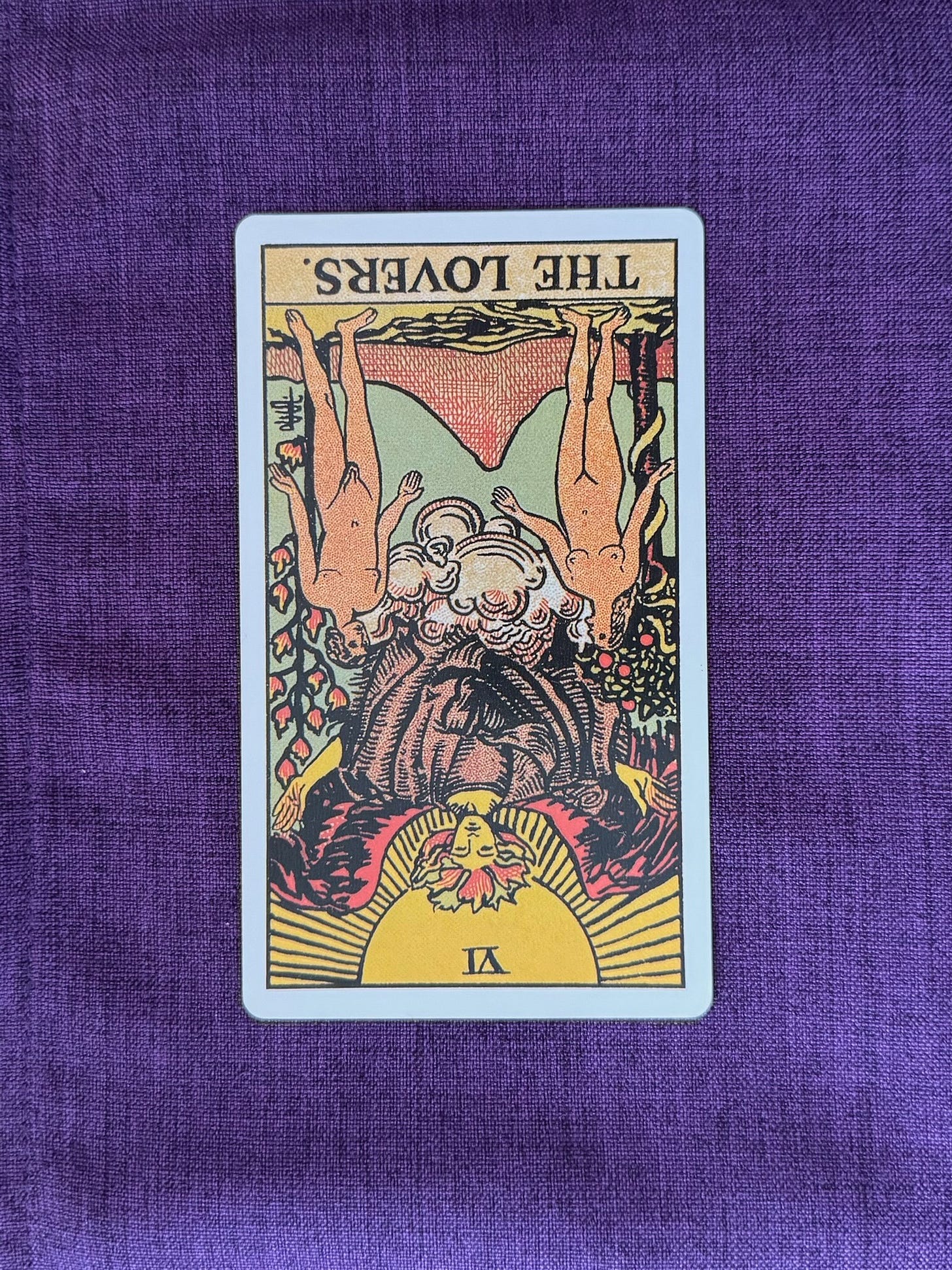 upside down Lovers card shows a naked man and woman standing beneath the angel, who blesses them. The couple stands in a beautiful, fertile landscape reminiscent of the Garden of Eden. Behind the woman stands a tall apple tree, with a snake winding its way up the trunk. Behind the man is a tree of 12 flames. Back and center is a volcanic mountain.