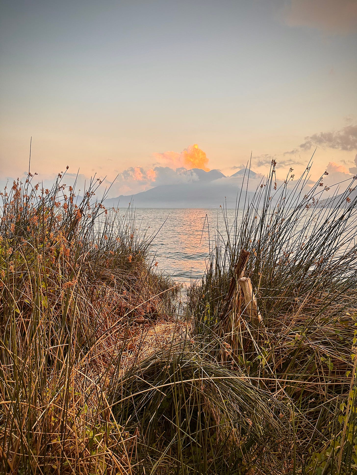 A calm lake is framed by volcanic peaks and green grasses in the soft evening light.