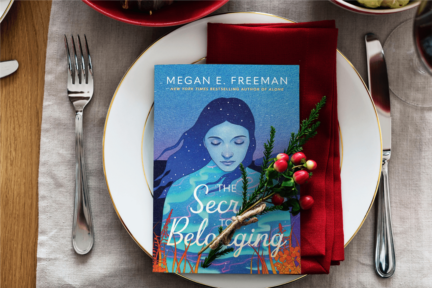 Book "The Secret to Belonging" on a plate with napkins, forks, knife and a stem of Holly rests on top of the book