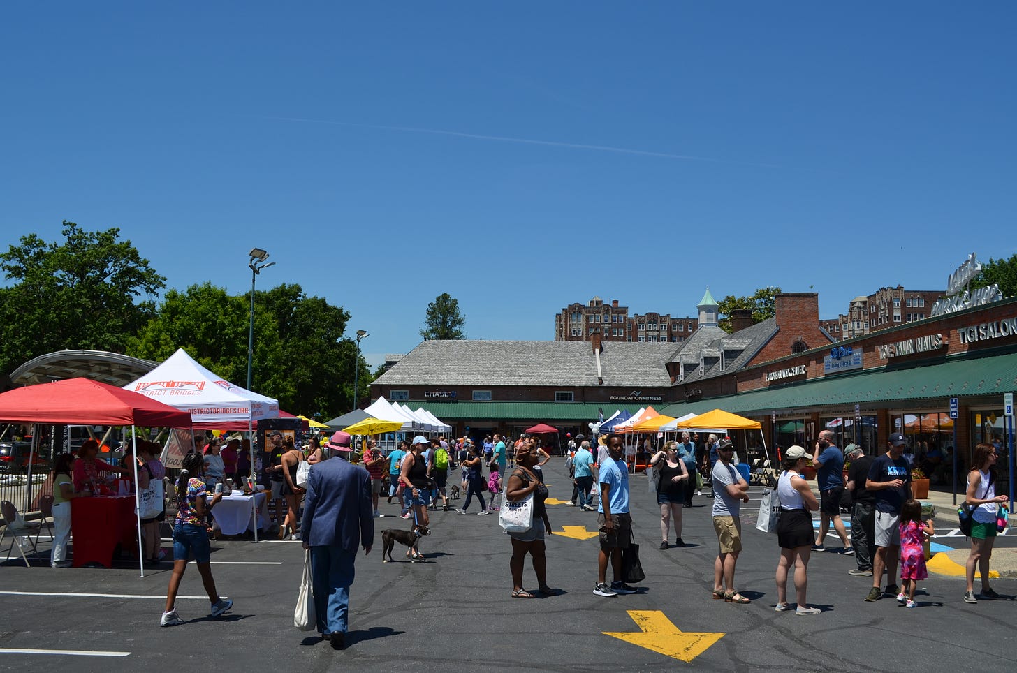Photo shows the cleveland park park and shop with tents and lots of people in the parking lot. The shops are in the background, the view is looking north. It's a clear day. There are about 50 people and children. There's lots of color in tent and clothes. The big yellow arrows in the parking lot ruin the view. It's a clear day and you can see the Broadmoor Apartments in the background.