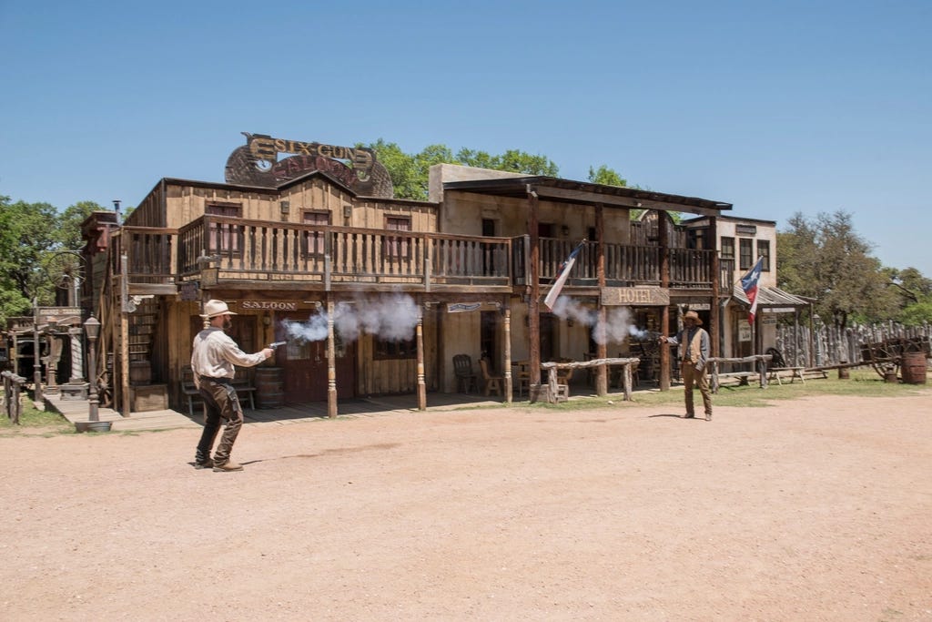 Two historical interpreters, standing in a gravel road outside a wooden building with a sign reading "Six-Gun Saloon" and shooting at each other with pistols.