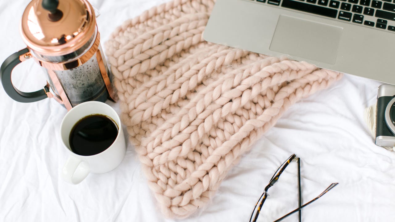 A cozy flat lay on a white bed featuring a chunky knit blanket, a French press and mug of coffee, a laptop, reading glasses, and a camera.