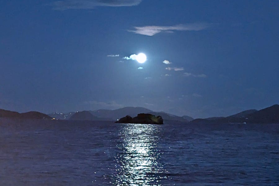 A bright Full Moon illuminates the evening sky over the ocean in St. Thomas, casting a shimmering reflection across the water. A small rocky island appears in the center of the scene, with distant hills silhouetted against the deep blue horizon. Soft clouds drift around the moon, creating a calm, serene atmosphere.