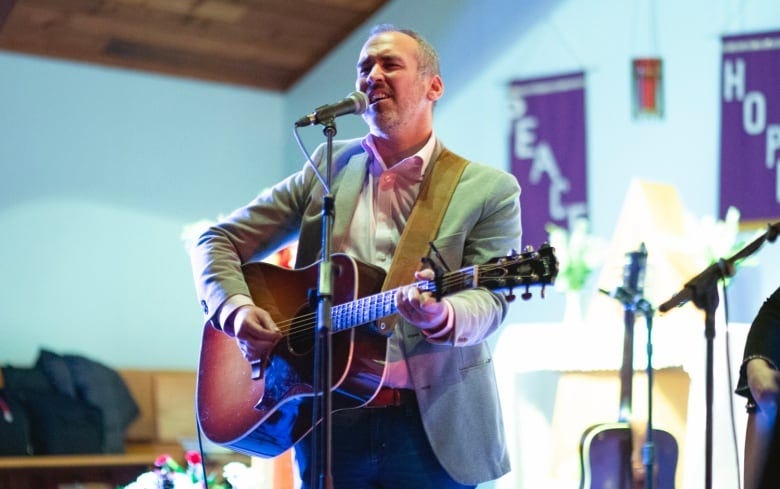 A man with a guitar stands behind a microphone. 