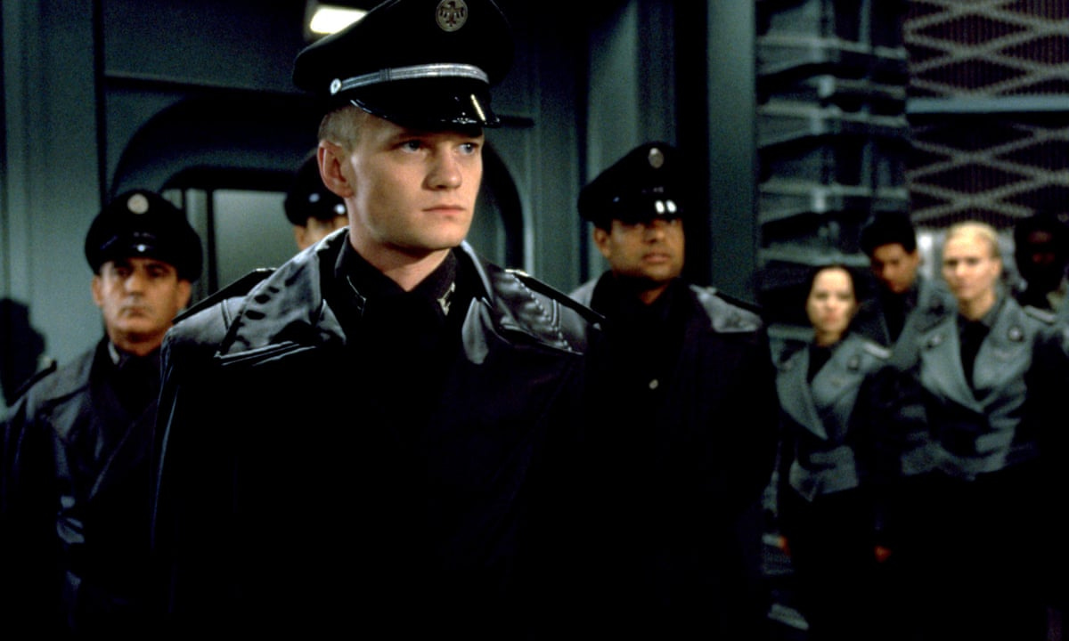 Soldiers in the nazi-like uniform, a tall white-haired man in the leather military jacket in front of the camera, a few officers behind him. A scene from the movie Starship Troopers.