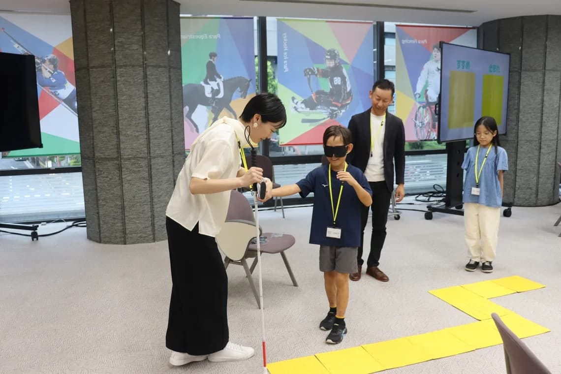 A blindfolded boy is guided by a volunteer holding a white cane, next to a path of yellow tactile paving tiles, while another student waits her turn.