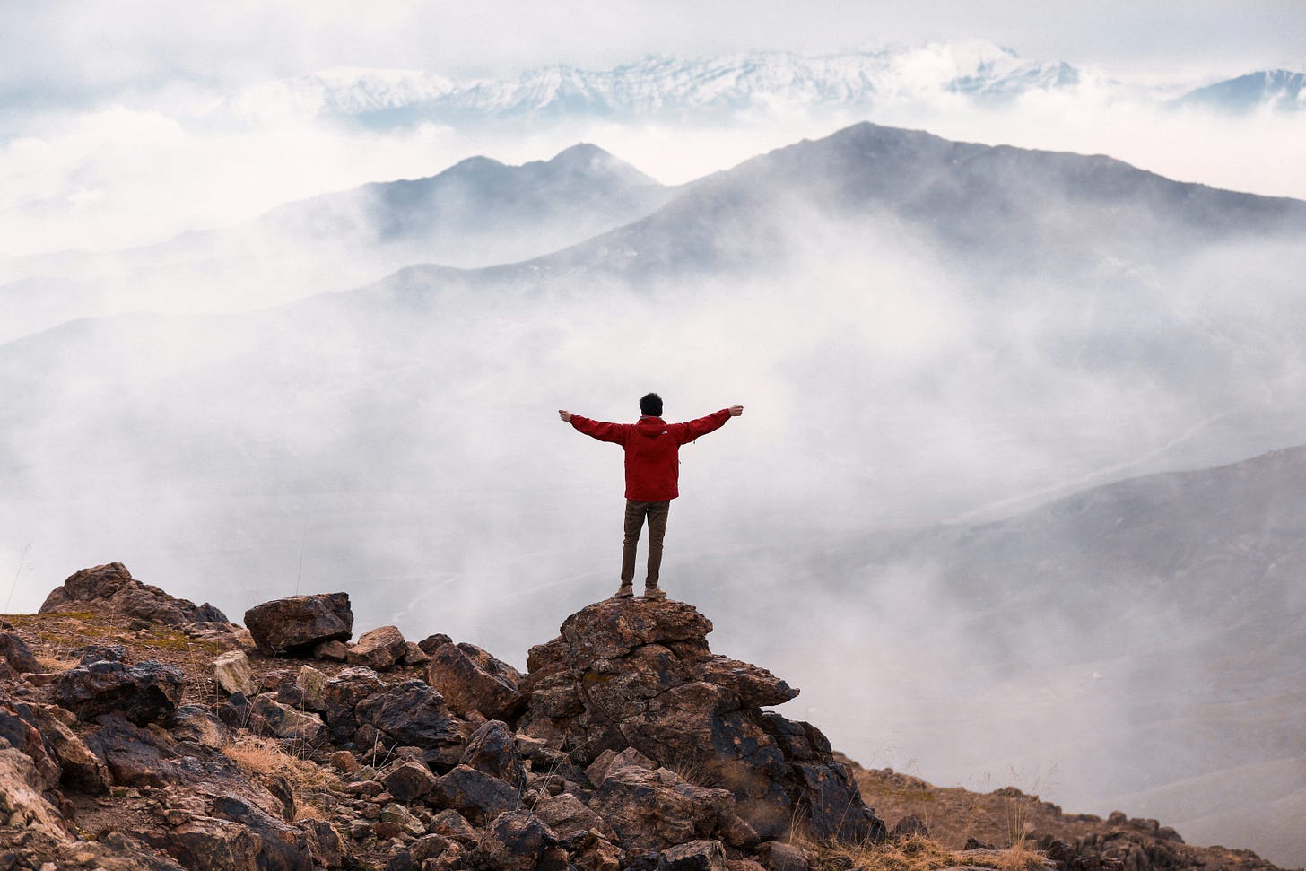 A man in a red jacket and black pants stands on a rock overlooking a mountain range. His arms are outstretched. There is a white foggy mist over the mountains.