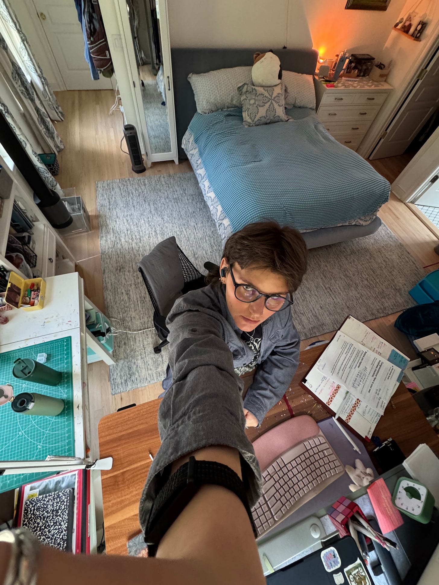 A zoomed-out selfie of me sitting at my desk. In front of me is a big pink keyboard and a planner filled with messy handwriting. Behind me is a bed, mirror, and dresser atop a light blue carpet.