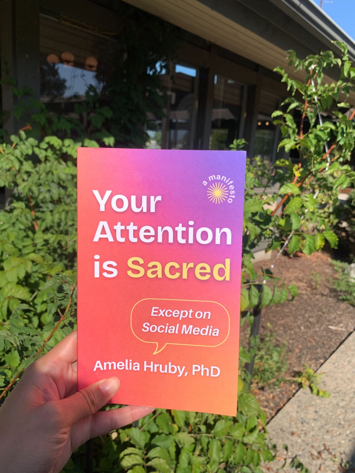 hand holding a book called your attention is sacred except on social media by amelia hruby phd in front of green foliage