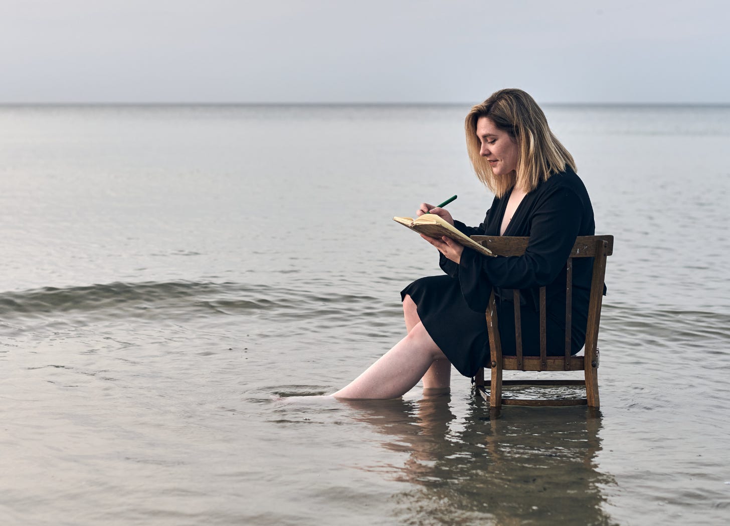 Contemplative woman writing in a journal while sitting in shallow water. Photo courtesy of TRAVELARIUM via Canva. 