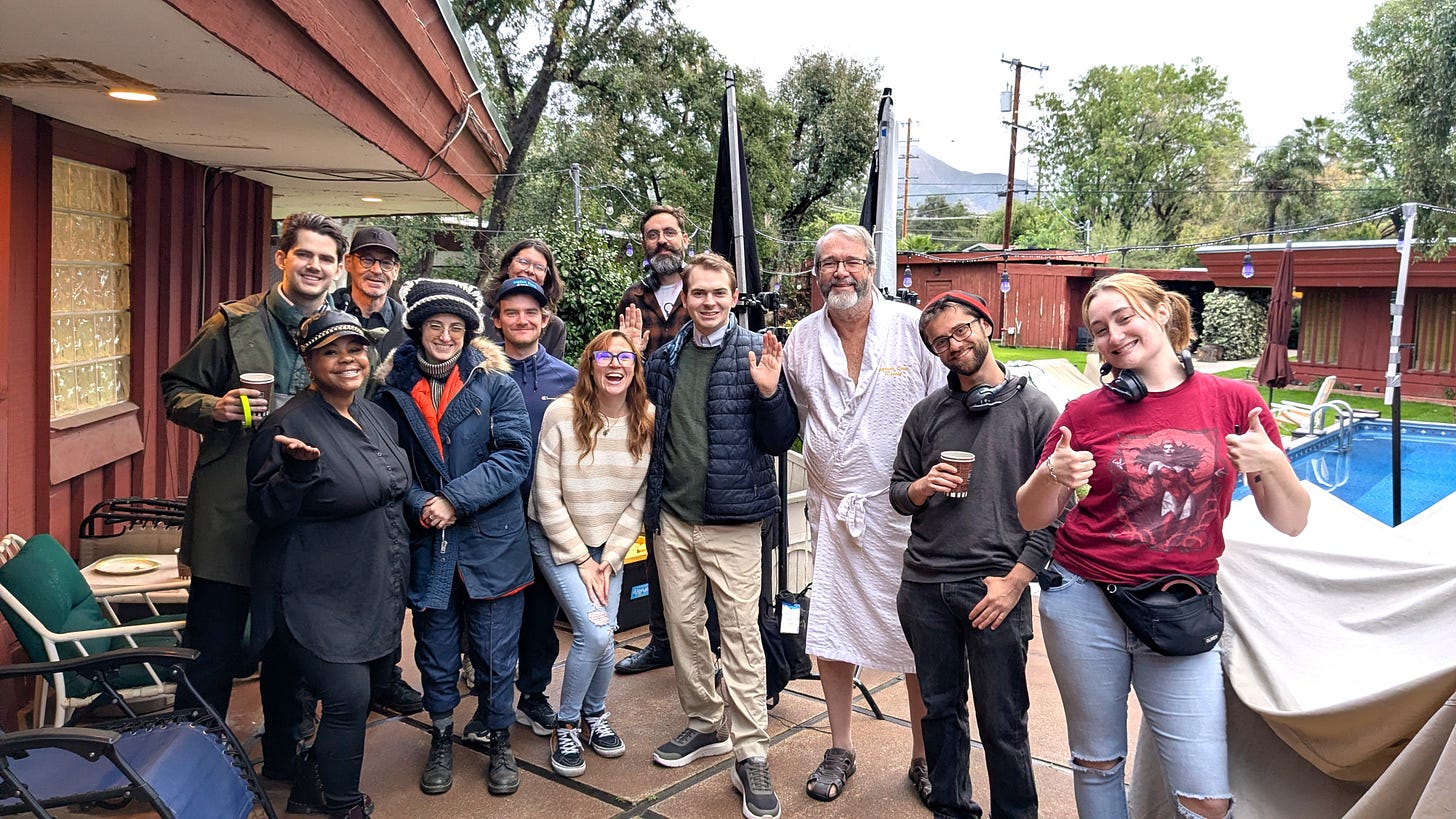 A group of about a dozen film crew members stand together outdoors on a patio set, smiling and posing for a behind-the-scenes photo. Some hold coffee cups, others give thumbs up or wave. The setting includes a mid-century style building, lighting stands, and a covered pool in the background, with trees and mountains visible beyond. One person near the center wears a white robe, while others are dressed casually in jackets and production attire, suggesting a cool-weather shoot day.