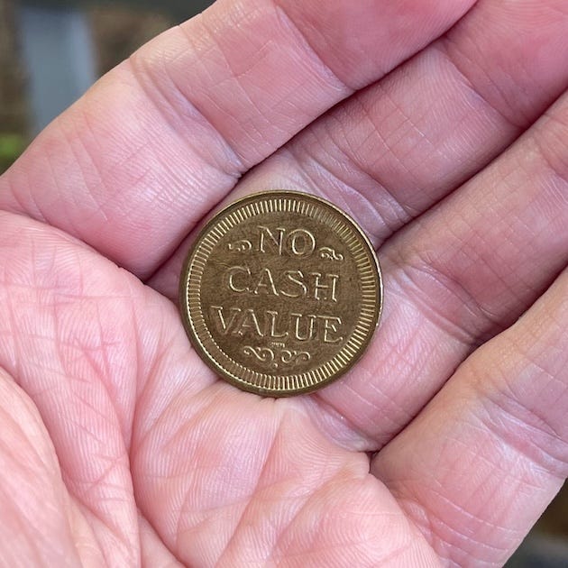 A close-up of a person’s open palm holding a small brass-colored token. The token reads “NO CASH VALUE” in raised capital letters. The coin is centered in the hand, lines and creases of the skin clearly visible, emphasizing texture and intimacy. The background is softly blurred, drawing attention to the object and gesture of holding.